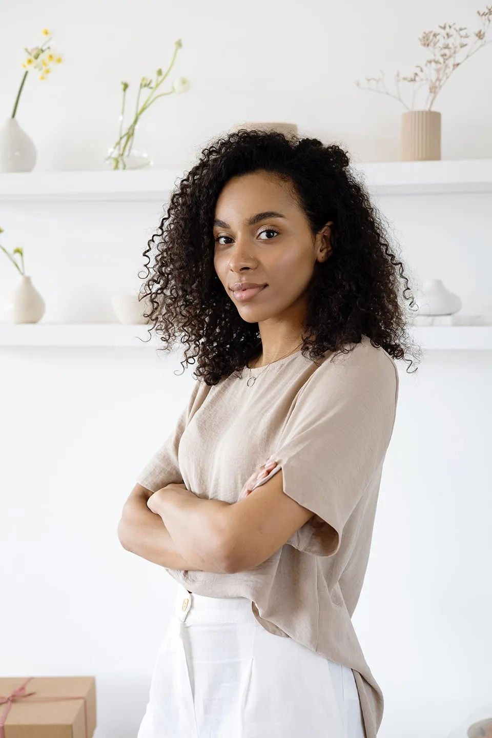 A designer with curly hair and folded arms in front of white shelves with vases and dried flowers.